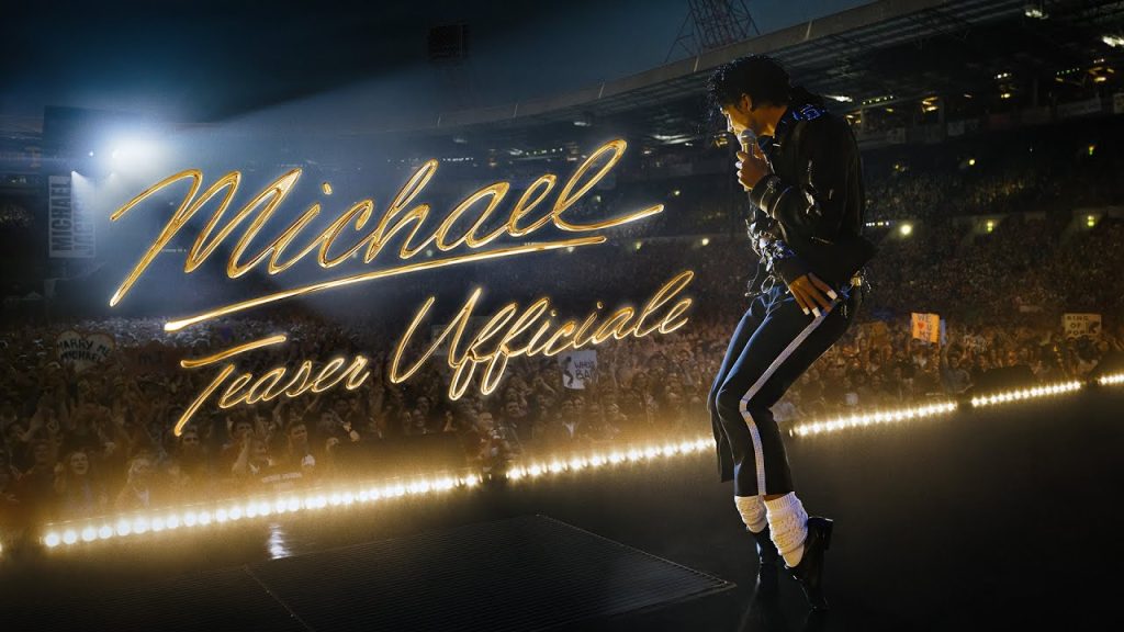 Male tribute performer on a darkened arena stage, singing into a microphone with a neon cursive backdrop reading 'Michael Teaser Unofficial' and a cheering, packed crowd behind him.