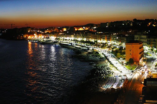 Pozzuoli - Passione Cinema Evening waterfront of a coastal town: sunset sky, bright streetlights along the harbor, boats docked by the quay, and calm water.