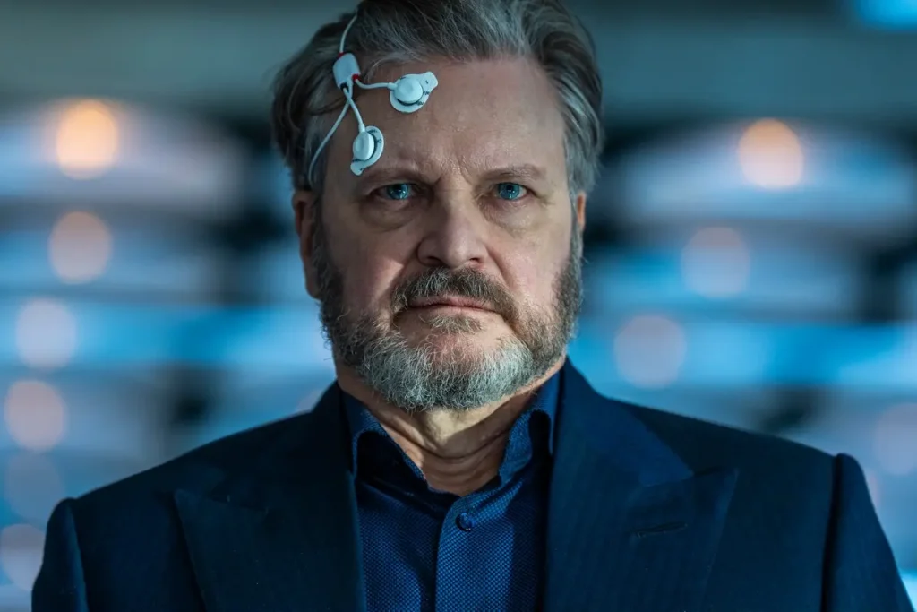 Older man in a suit with EEG electrodes attached to his head, looking directly at the camera in a clinical setting.