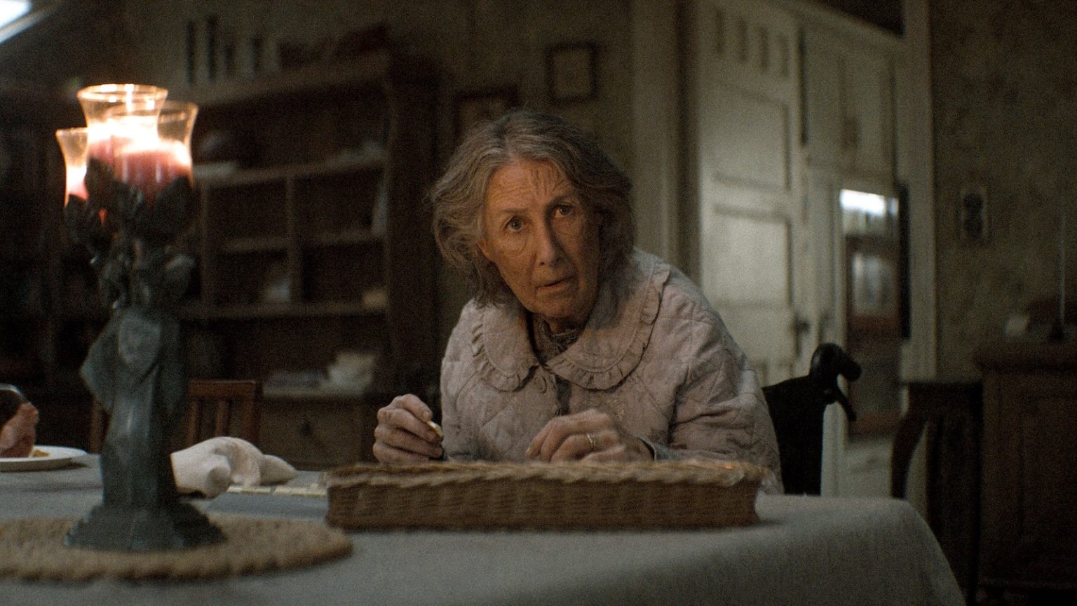 Older woman with gray hair sits at a table and prepares a pie in a dim, candlelit kitchen.